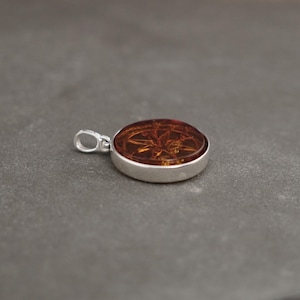 Op de afbeelding: Een ronde amber hanger met een zilveren zetting. De amber heeft een gedetailleerd ontwerp aan de binnenkant.