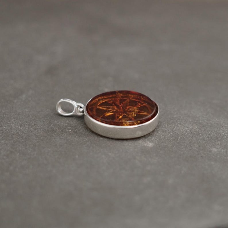 Op de afbeelding: Een ronde amber hanger met een zilveren zetting. De amber heeft een gedetailleerd ontwerp aan de binnenkant.