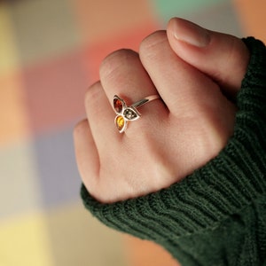 Op de afbeelding: Een zilveren ring met drie amberstenen gezet in een bladerenontwerp. De ring wordt gedragen op een hand met een groene trui.