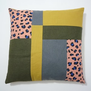 May include: A square pillow with a patchwork design. The pillow is made of four different fabrics: a pink and blue leopard print, a solid yellow fabric, a solid dark green fabric, and a solid gray fabric.