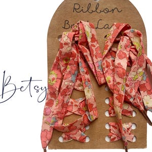 May include: A pair of coral-toned floral fabric shoelaces with metallic aglets, mounted on a brown cardboard display card labeled Liberty Tana Lawn and Betsy. The laces feature a dense pattern of small pink, red, and yellow flowers on a light background.
