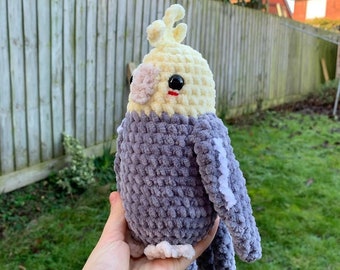 May include: A handmade, plush cockatiel bird in shades of gray, cream, and yellow. The bird has black eyes, a small red beak, and a crest. The soft, textured bird is a cuddly toy.