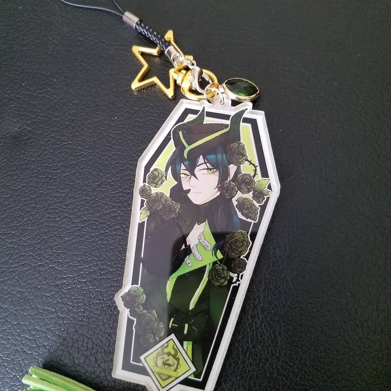 May include: A black and green character with horns and a green hat is depicted in a coffin-shaped acrylic charm. The character is surrounded by black roses and has a green and black outfit. The charm is attached to a black string with a gold star and a green gem.