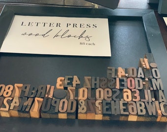 May include: A collection of wooden letterpress blocks in various sizes and colors, including letters, numbers, and symbols. The blocks are arranged in a random pattern and are displayed in a black frame. A sign above the blocks reads "LETTER PRESS wood blocks $8 each".
