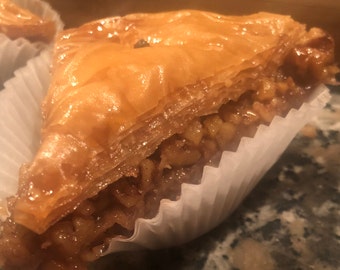 May include: A single serving of baklava, a traditional Greek pastry, in a white paper cup. The baklava is made with layers of phyllo dough, filled with chopped nuts, and drizzled with honey syrup.
