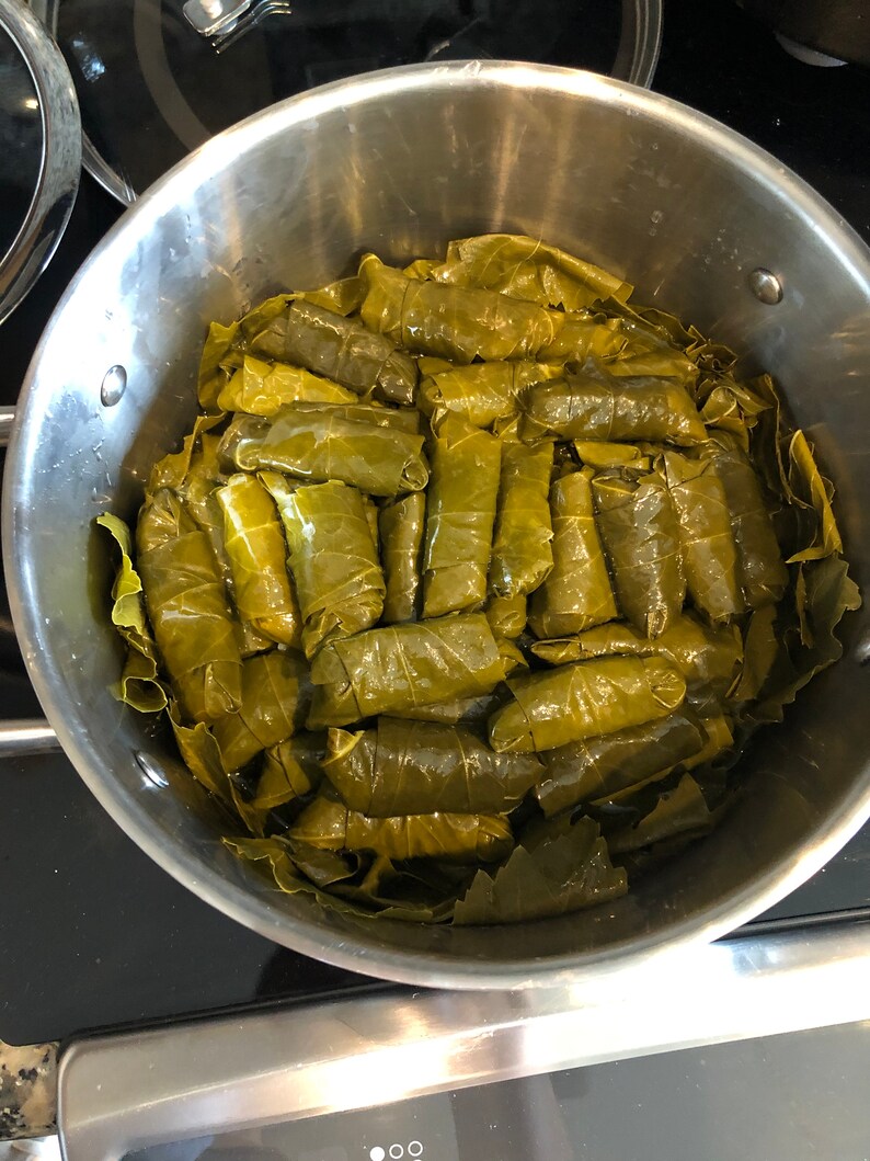 Pu&ograve; includere: Una pentola in acciaio inox piena di una grande quantit&agrave; di foglie di vite verdi avvolte attorno a un ripieno, probabilmente per fare dolmades.