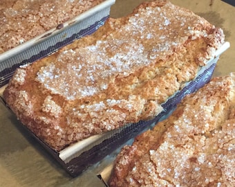 May include: Three loaves of bread with a sugary crust, baked in brown paper-lined loaf pans. The bread is golden brown and has a slightly rough texture.