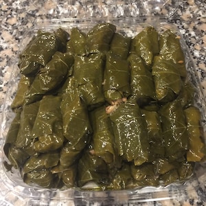 Pu&ograve; includere: Un contenitore di plastica trasparente pieno di una grande quantit&agrave; di foglie di vite verdi avvolte attorno a un ripieno, probabilmente riso e carne, formando un piatto tradizionale chiamato dolmades.