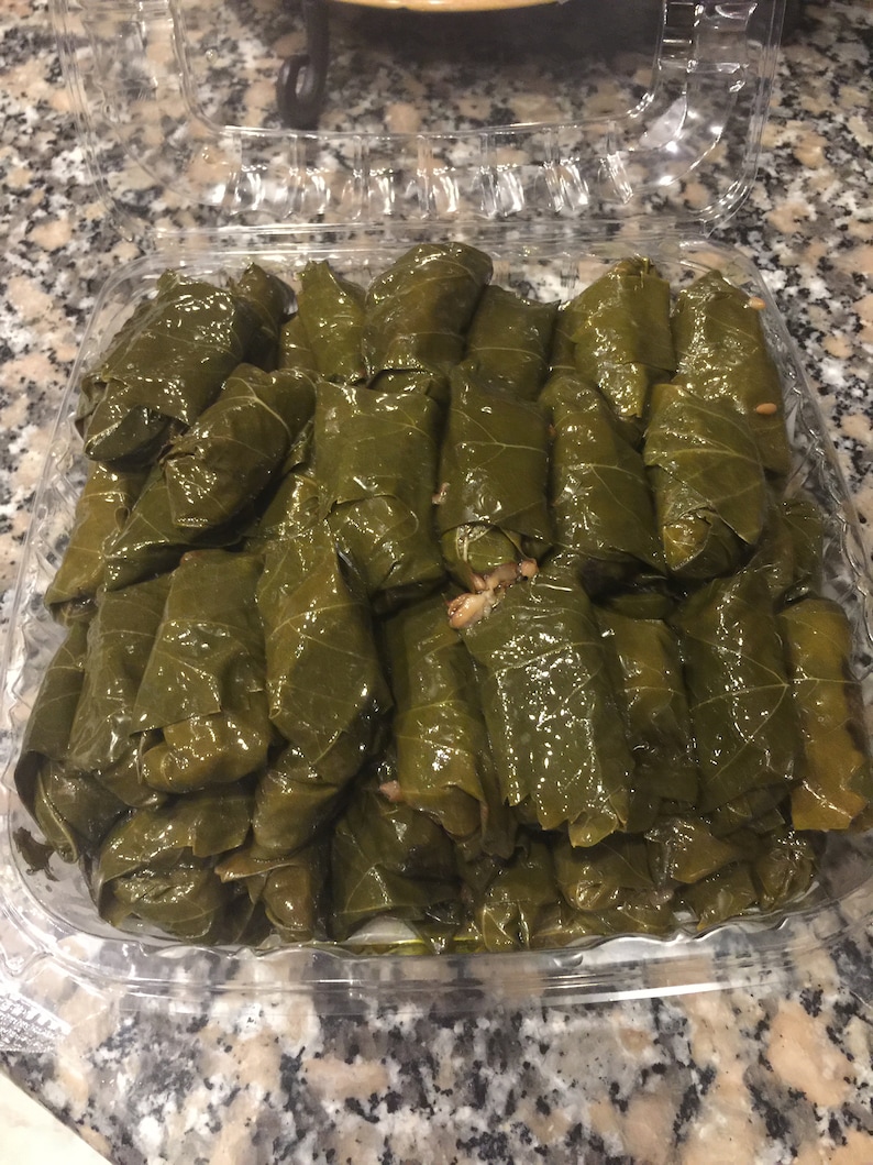 Pu&ograve; includere: Un contenitore di plastica trasparente pieno di una grande quantit&agrave; di foglie di vite verdi avvolte attorno a un ripieno, probabilmente riso e carne, formando un piatto tradizionale chiamato dolmades.