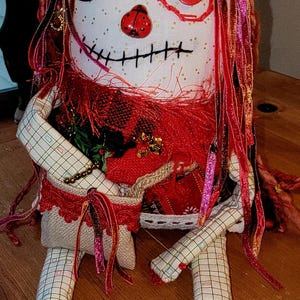 May include: A handmade rag doll with a whimsical design. The doll features a white face with a stitched mouth, a large black button eye, and a red button eye. It has red yarn hair and is dressed in a patchwork outfit with red accents.
