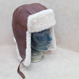 May include: A brown leather aviator hat with a white faux fur trim. The hat has a long strap that ties under the chin.