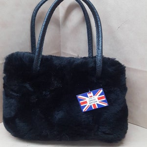 May include: A black faux fur handbag with a black leather handle. The bag has a small tag attached to it that says "Made in Great Britain".