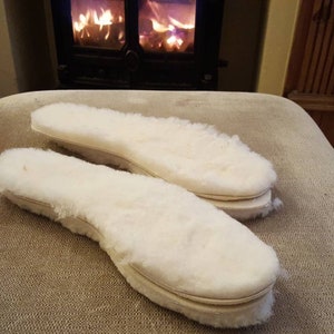 May include: Two white sheepskin insoles with a brown leather trim. The insoles are lying on a beige surface.