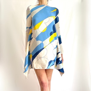 May include: A short dress with a colorful abstract pattern. The dress features a design of blue, yellow, and white shapes. The model is wearing silver-colored high heels. The dress has long sleeves and a unique, asymmetrical hemline.
