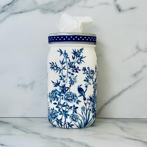 May include: A white ceramic jar with a blue and white floral design and a blue polka dot lid. The jar is filled with tissues and is sitting on a white marble surface.