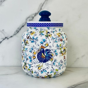 May include: White ceramic jar with a blue lid and a rooster design. The jar is decorated with floral patterns in blue, yellow, and green.