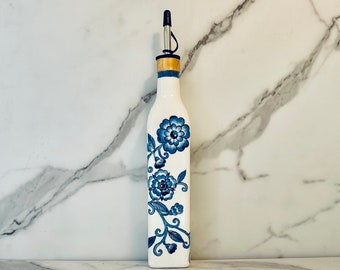 May include: White ceramic olive oil bottle with a blue floral design. The bottle has a black pour spout and a gold colored top.