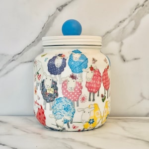 May include: A white jar with a lid and a blue knob. The jar is decorated with colorful sheep illustrations. The sheep have various patterns and colors, including blue, pink, and yellow. The jar is on a white marble surface.