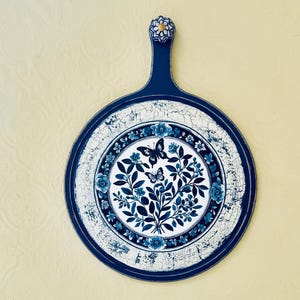 May include: Decorative blue and white round cutting board with a handle. The board features a floral and butterfly design in the center, surrounded by a white and blue border. The handle is topped with a decorative knob.
