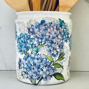 May include: A cylindrical ceramic utensil holder featuring a white base decorated with a vibrant pattern of blue and purple hydrangea flower clusters, green leaves, and small butterflies, containing several light brown wooden kitchen utensils.
