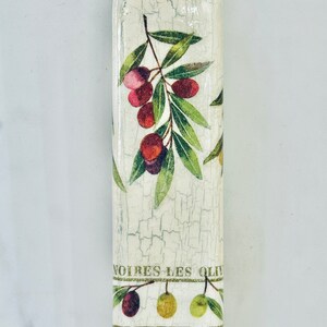 Puede incluir: Una botella blanca con un acabado desgastado y un dise&ntilde;o de ramas de olivo con hojas verdes y aceitunas rojas. La botella tiene el texto "NOIRES LES OLIV" y "OURNANTES LES" impreso en ella.