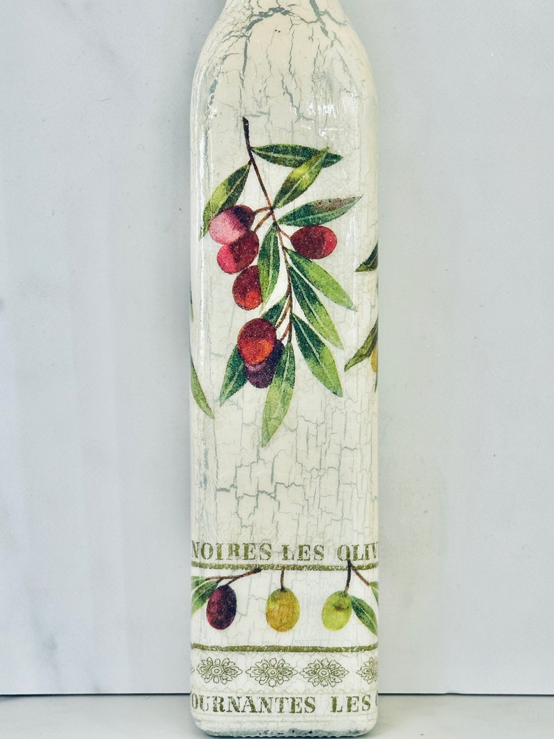Puede incluir: Una botella blanca con un acabado desgastado y un dise&ntilde;o de ramas de olivo con hojas verdes y aceitunas rojas. La botella tiene el texto "NOIRES LES OLIV" y "OURNANTES LES" impreso en ella.