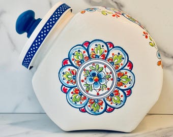 May include: A white ceramic canister with a blue knob lid featuring a blue and white polka dot rim. The body is decorated with a large, hand-painted floral mandala pattern in shades of blue, green, orange, and red, centered on the side.