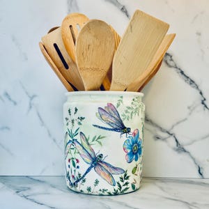 May include: A white ceramic utensil holder with a dragonfly and floral design. The holder is filled with wooden spoons and spatulas.
