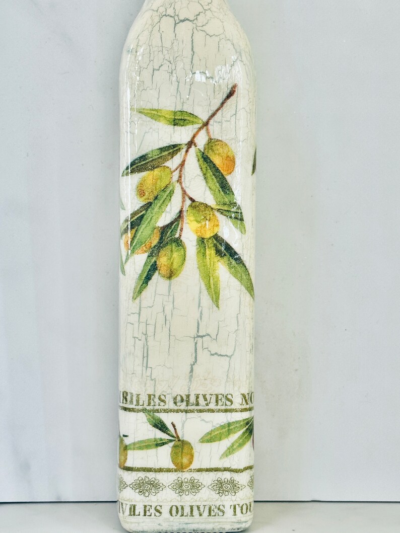 Puede incluir: Una botella blanca con un acabado desgastado presenta un dise&ntilde;o de ramas de olivo con hojas verdes y aceitunas amarillas. La botella tiene el texto "ILES OLIVES NO" y "ILES OLIVES TO" impreso en ella.