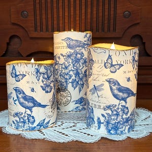 May include: Three white pillar candles with blue and white floral designs and text. The candles have a vintage aesthetic and feature images of birds, butterflies, and dragonflies.