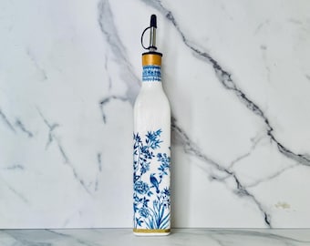 May include: White glass oil bottle with a blue floral design. The bottle has a stainless steel spout and a wooden and blue patterned collar. The floral design includes a bird and various plants. The base has a gold band.