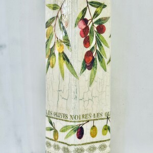 Puede incluir: Una botella blanca con un acabado desgastado y un dise&ntilde;o de ramas de olivo y aceitunas. La botella tiene el texto "Les Olives Noires Les Ol" y "Les Olives Tournantes Le" impreso en ella.