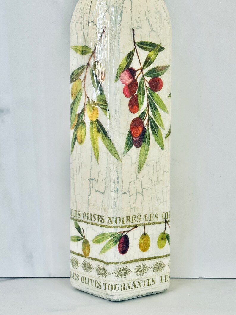 Puede incluir: Una botella blanca con un acabado desgastado y un dise&ntilde;o de ramas de olivo y aceitunas. La botella tiene el texto "Les Olives Noires Les Ol" y "Les Olives Tournantes Le" impreso en ella.