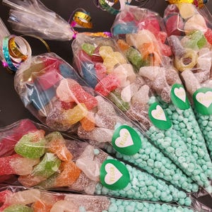May include: Five clear plastic cone-shaped bags filled with assorted colorful gummy candies and topped with light blue candy beads. Each bag features a green heart-shaped sticker with the word VEGAN printed in white and a metallic ribbon tie.
