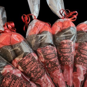 Puede incluir: Varias bolsas de celofán llenas de caramelos rojos y negros, atadas con cintas rojas. Algunas bolsas tienen etiquetas de "Stranger Things", otras dicen "Happy Birthday". Los caramelos son de diferentes formas y tamaños, sobre un fondo oscuro.