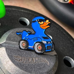 May include: A blue duck wearing sunglasses and a racing suit with the text "ZED" on the side. The duck is holding a steering wheel and is sitting on a silver metal surface with the text "DUCK WHEELS" printed on it. The duck is a pin and is part of the "DUCK WHEELS PATCH SERIES".