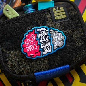 May include: A black tactical pouch with a hook-and-loop front panel featuring a colorful embroidered patch that reads "GOOD DAYS FIGHT FOR EVERY DAY BAD DAYS". The pouch contains a yellow rubber duck, a small green tool, and a blue metal item.