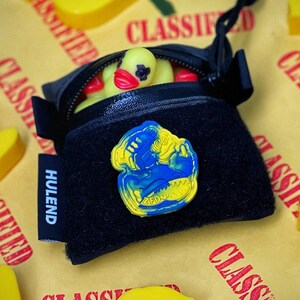 May include: A black zippered pouch with a HULEND tag and a blue and yellow rubber patch on the front. The pouch contains a small yellow rubber duck with a red beak and black eye markings. The pouch is surrounded by several yellow rubber ducks.
