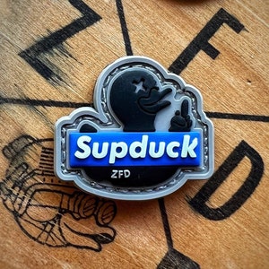 May include: A silver and blue patch with a black rubber duck design. The patch has the text "Supduck" in blue letters and "ZFD" in white letters.