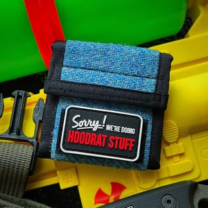 May include: A small blue and black wallet with a white patch that says "Sorry! We're doing Hoodrat Stuff". The wallet is sitting on top of a yellow and black toy gun.