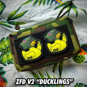 May include: A camouflage zippered pouch with two yellow rubber ducky patches. The duckies are wearing military helmets with American flags on them. The text "ZFD V2 "DUCKLINGS"" is printed below the pouch.