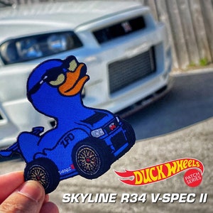 May include: An embroidered patch featuring a blue rubber duck character wearing sunglasses, designed to resemble a Skyline R34 V-Spec II car with wheels, the text ZFD, and a red GT emblem. The patch includes the Duck Wheels logo and text.