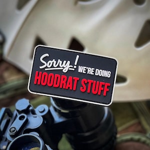 May include: A rectangular black morale patch with a white border featuring the white text Sorry! WE'RE DOING and the red text HOODRAT STUFF. The patch is positioned in front of a tan tactical helmet and black optical equipment with a circular logo.