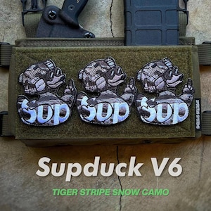 May include: A green tactical vest with three patches featuring a cartoon duck wearing sunglasses and the word "Sup". The vest has two black straps with buckles and a black magazine pouch. A black knife with a gold "Defcon" logo is attached to the vest.
