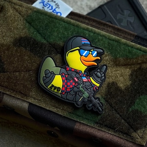 May include: A yellow rubber duck wearing a red and black plaid shirt, blue sunglasses, and a black hat with a US flag design. The duck is holding a black rifle and has the letters "ZFD" on its side. The duck is attached to a camouflage fabric pouch with the words "Spiritus Systems" printed on it.