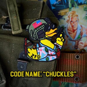 May include: A yellow rubber ducky wearing a blue and pink Hawaiian shirt, a black helmet with an American flag, and goggles. The ducky is holding a gun in its right hand and has the text "ZFD" on its chest. The text "CODE NAME: "CHUCKLES"" is below the ducky.