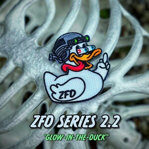 May include: A white embroidered patch featuring a cartoon duck wearing a tactical helmet with night vision goggles, a red tongue, and a raised wing. The duck has the text ZFD printed on its side. The patch is displayed over a white rib cage structure.
