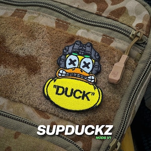 May include: A yellow embroidered patch with the word "DUCK" in black letters. The patch features a cartoon duck with an angry expression, wearing a black helmet with green accents and X-shaped eyes. The patch is attached to a brown camouflage fabric.