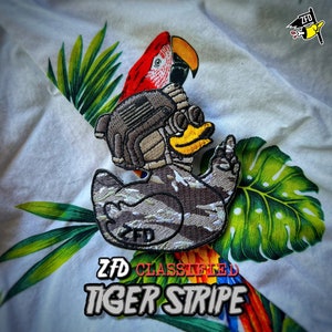 May include: An embroidered patch featuring a yellow rubber duck wearing a tactical helmet with night vision goggles and a camouflage pattern body. The duck is making a hand gesture. The text ZFD CLASSIFIED TIGER STRIPE is printed below.