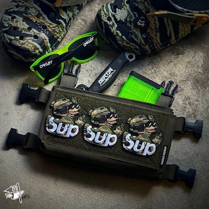 May include: A green tactical utility pouch with three patches that say "Sup" in white letters. The pouch has black straps and buckles. A green magazine, a black knife, and a pair of green sunglasses are visible in the background.